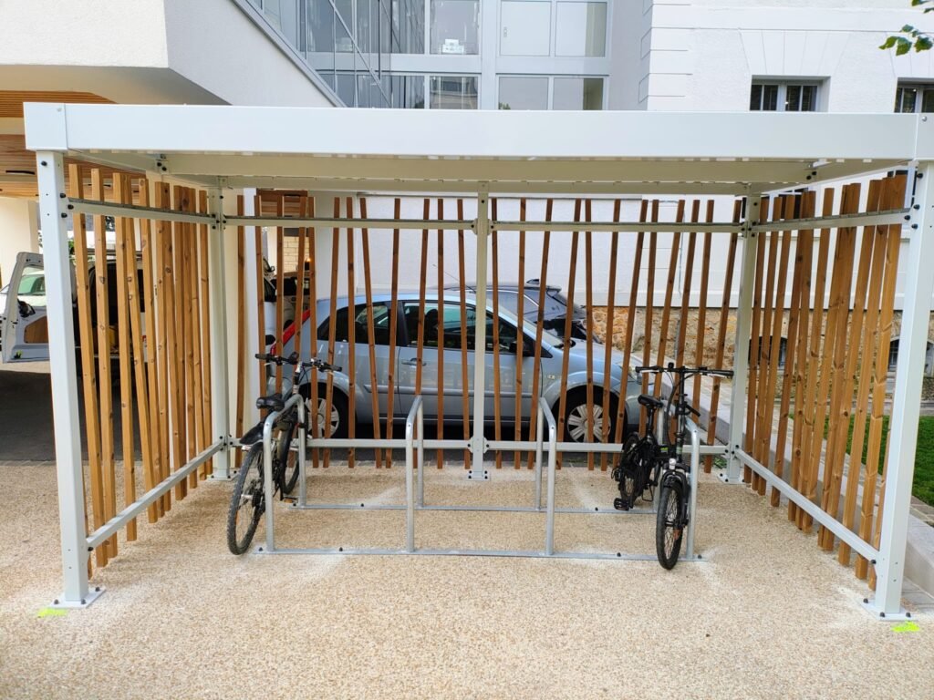 Abri vélo design avec structure blanche et parois en lames de bois, équipé d’arceaux de stationnement, contenant deux vélos garés.