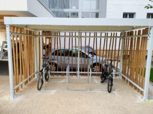 Abri vélo design avec structure blanche et parois en lames de bois, équipé d’arceaux de stationnement, contenant deux vélos garés.