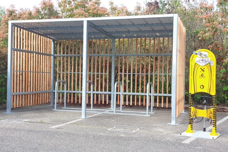Wooden bicycle shelter designed by ByCommute with a yellow inflation station on the front. The shelter features a metal structure and natural wood panels, providing a secure space for bicycle parking. The air terminal allows cyclists to easily inflate their tires. The whole unit is installed on asphalt, surrounded by vegetation.