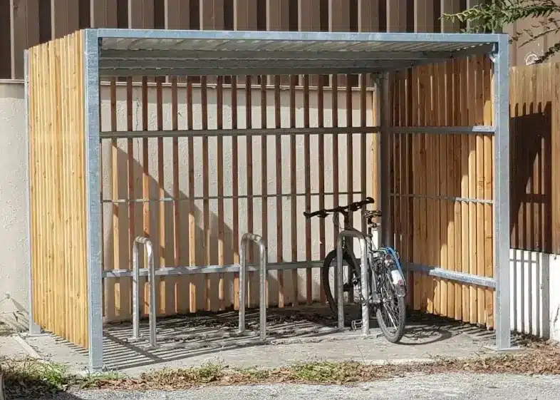 Cobertizo abierto para bicicletas con estructura metálica y paredes de listones de madera, que contiene varias bicicletas.