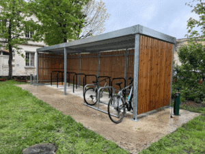 Abri vélo bois fermé avec structure métallique, équipé d’arceaux de stationnement et situé dans un espace vert urbain