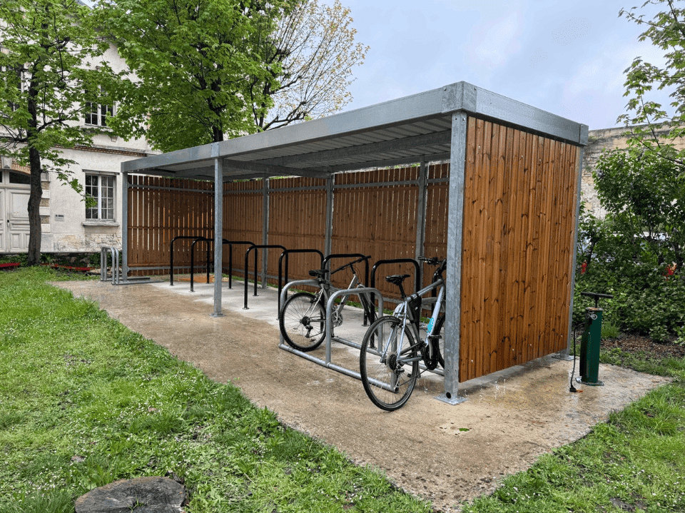 Abri vélo bois fermé avec structure métallique, équipé d’arceaux de stationnement et situé dans un espace vert urbain