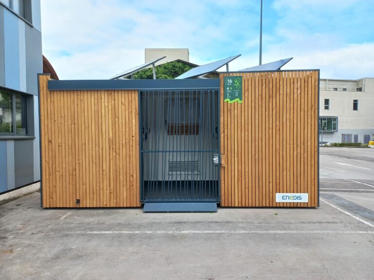 Garaje cerrado de madera para bicicletas con puerta de malla metálica, equipado con paneles solares en el techo, instalado en un aparcamiento de empresa.