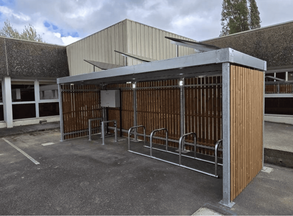 Abri vélo design en bois avec structure métallique galvanisée, rack à vélos vide, installé dans une cour d'entreprise sous un ciel partiellement nuageux.