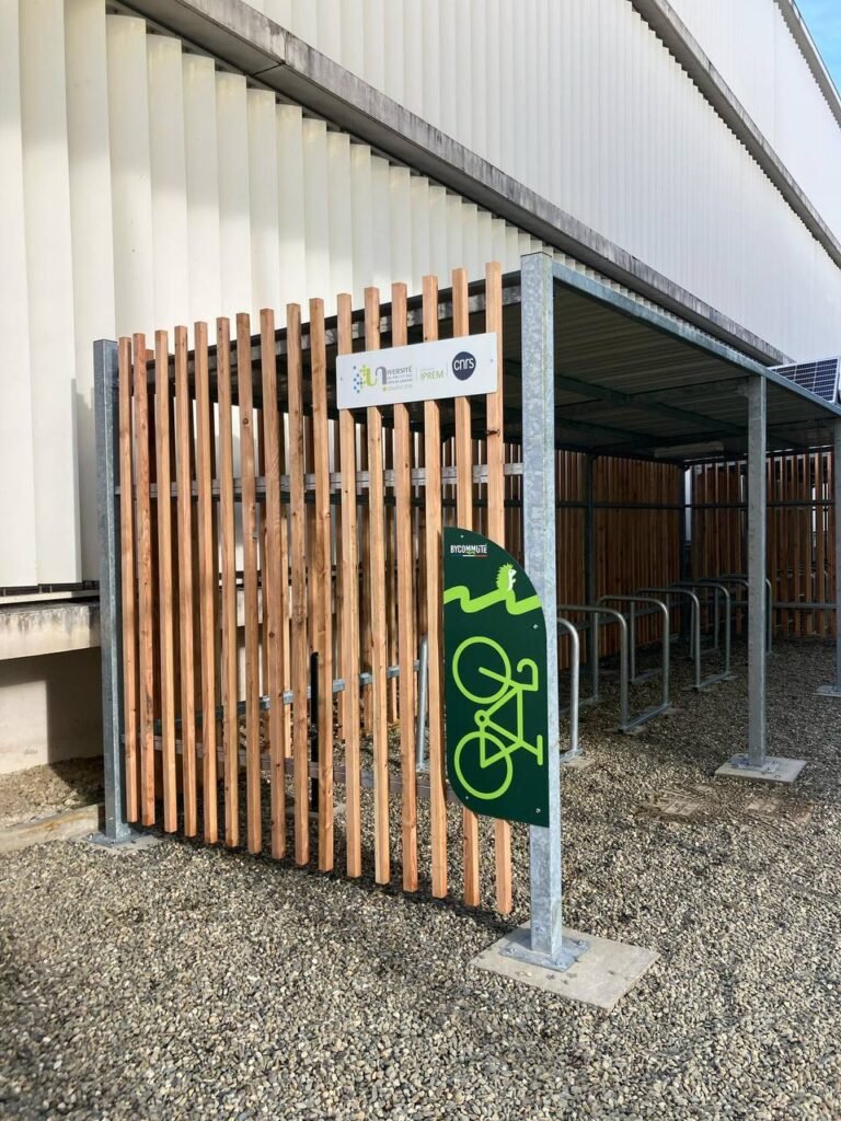 Abri vélo collectivité avec structure métallique et parois en bois, installé devant un bâtiment universitaire, équipé de racks vélos et signalétique Bycommute.