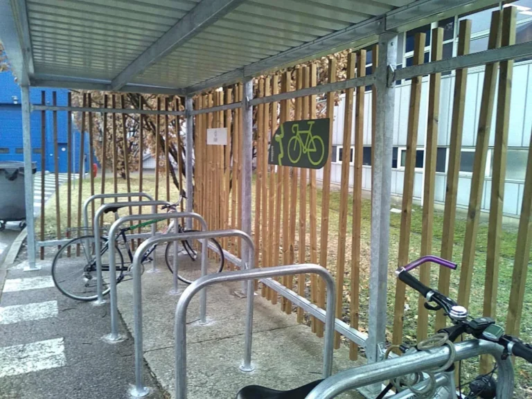 Abri vélo entreprise en métal avec arceaux de stationnement sous toit, installé près d'un bâtiment et protégé par une clôture en bois vertical.