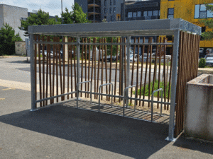 Abri vélo design "Jungle" avec structure métallique et lames verticales en bois, installé sur un parking en zone urbaine.