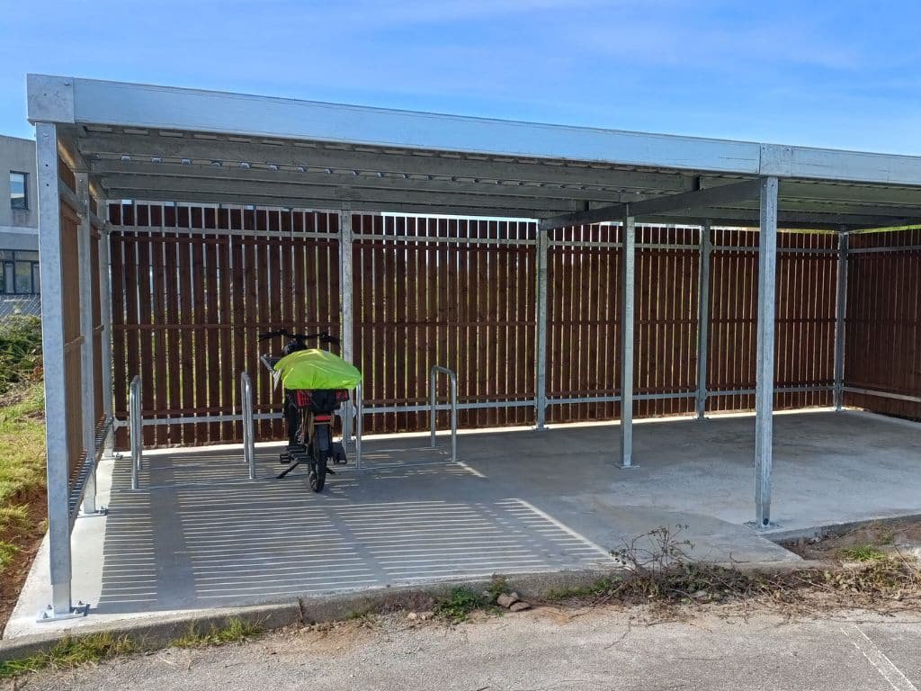 Abri vélo bois avec structure métallique, équipé d’arceaux de stationnement et abritant un vélo recouvert d’une housse verte.