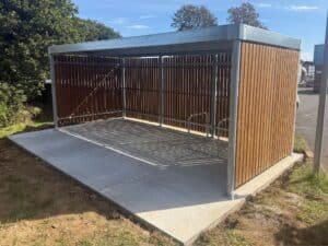 Abri vélo bois design avec structure métallique galvanisée et parois en lames de bois, installé sur une dalle béton dans un espace vert.